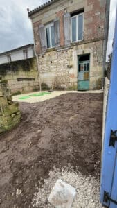 Maison en pierre en rénovation, cour boueuse avec gravier et couvercles verts de fosse septique.
