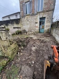 Terrassement devant une maison ancienne en pierre en cours de rénovation avec porte turquoise et engin de chantier.